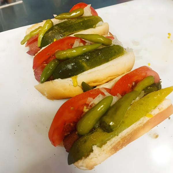 Three hot dogs in buns topped with tomatoes, pickles, and green peppers on a white surface.